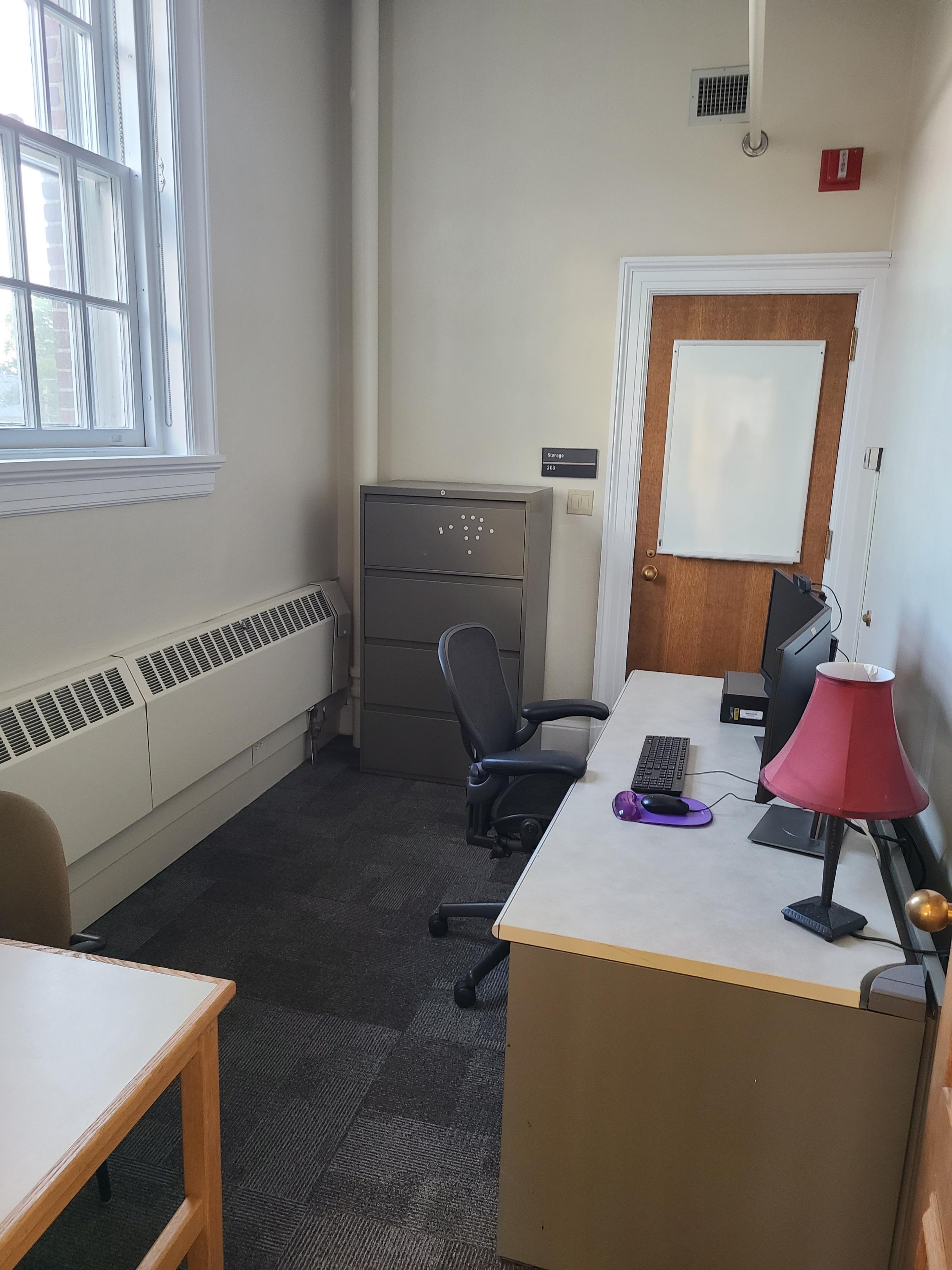 Office with large window, a single desk with two monitors at a PC workstation, lamp, comfortable chair and second table and chair.