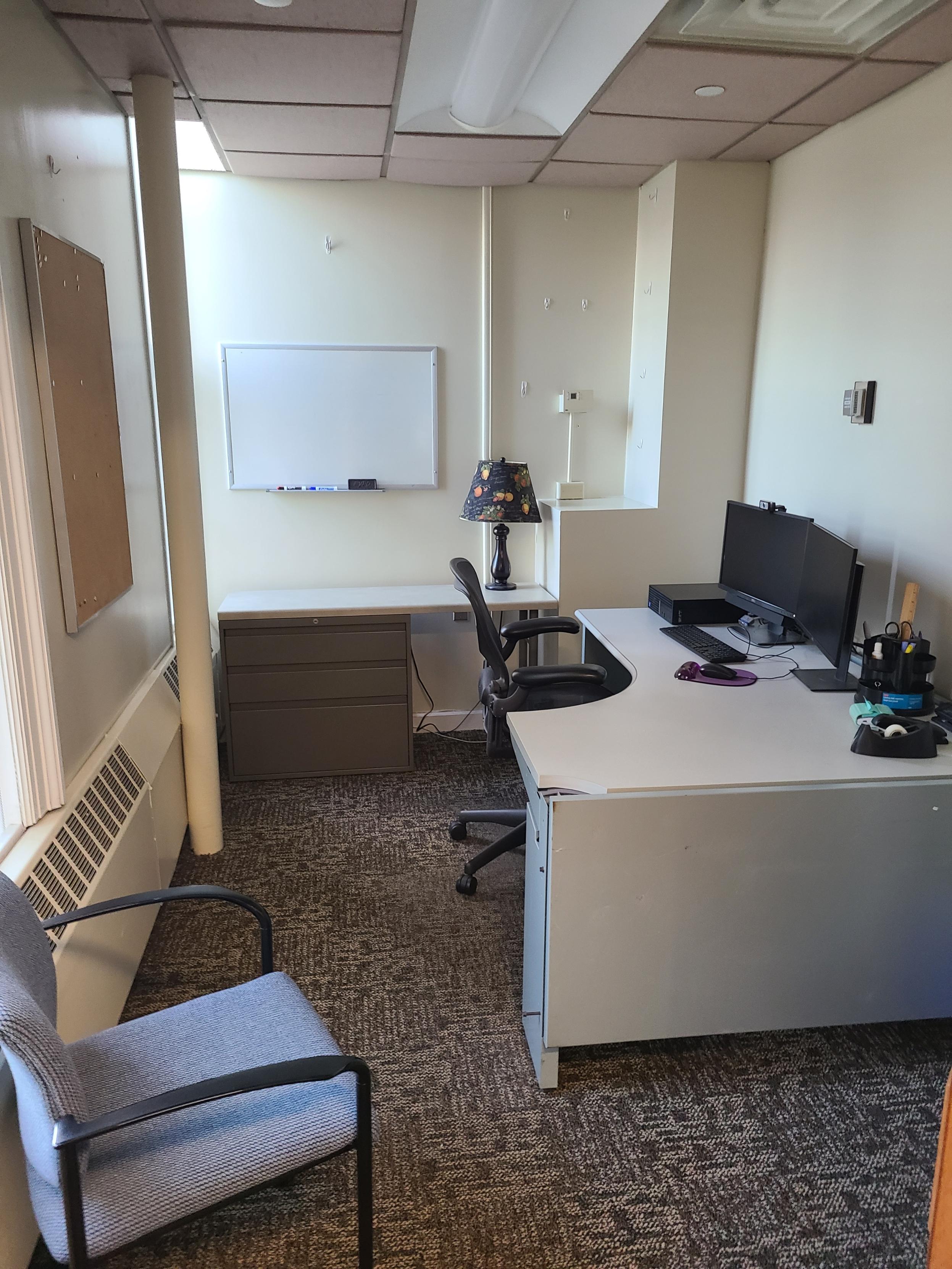 Office with a small corner desk, a PC workstation with two monitors, a comfortable chair and second side chair. Natural light comes in from two windows on either side of a bulletin board on the wall behind the desk. A whiteboard hangs on a wall over another small desk with a lamp and filing cabinet underneath.