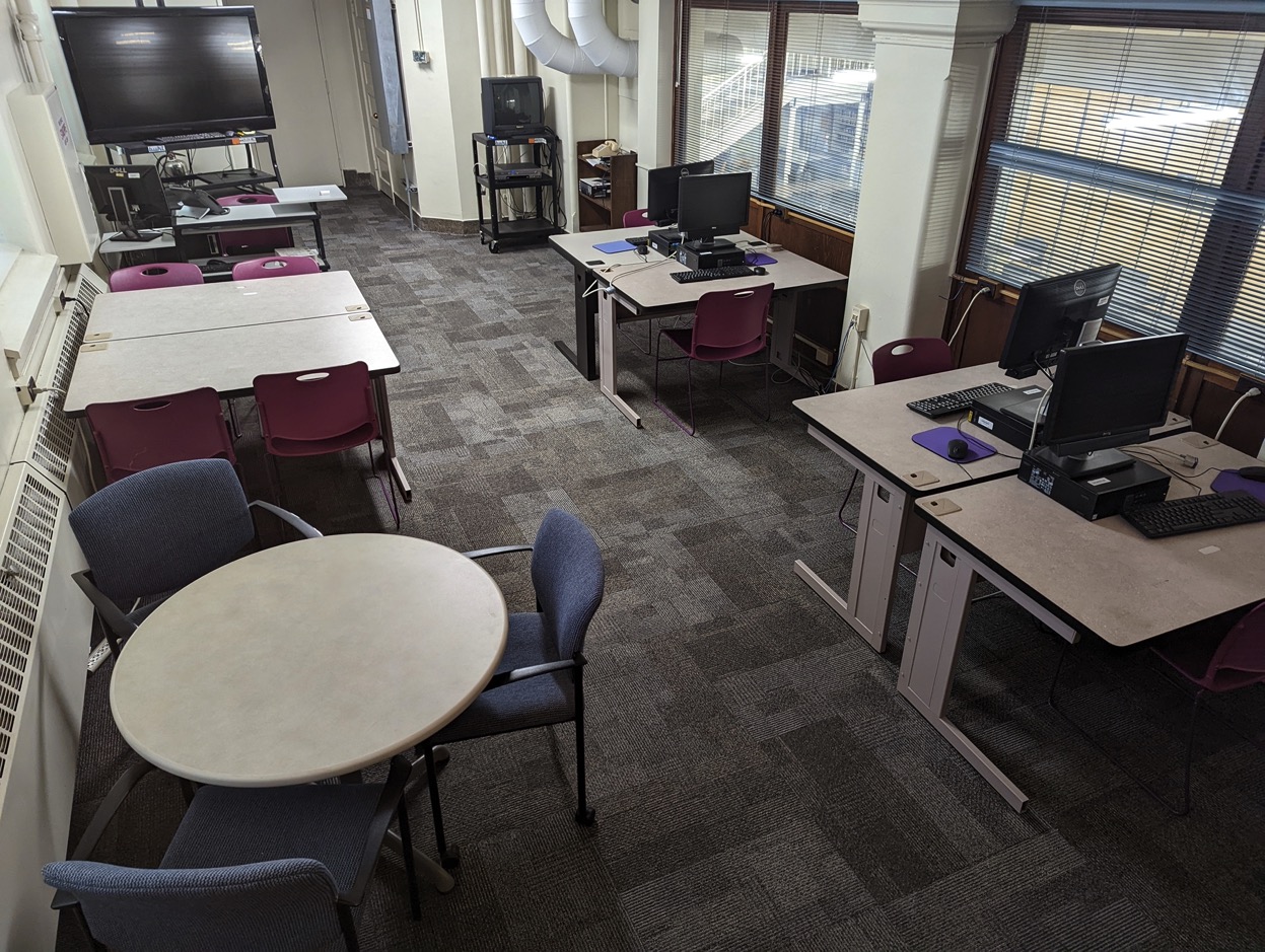 Bird's eye view of classroom with a large television on a rolling cart, a small television on a rolling cart with attached vcr, one square table with 4 chairs, one round table with 3 chairs, and 4 individual workstations with computers